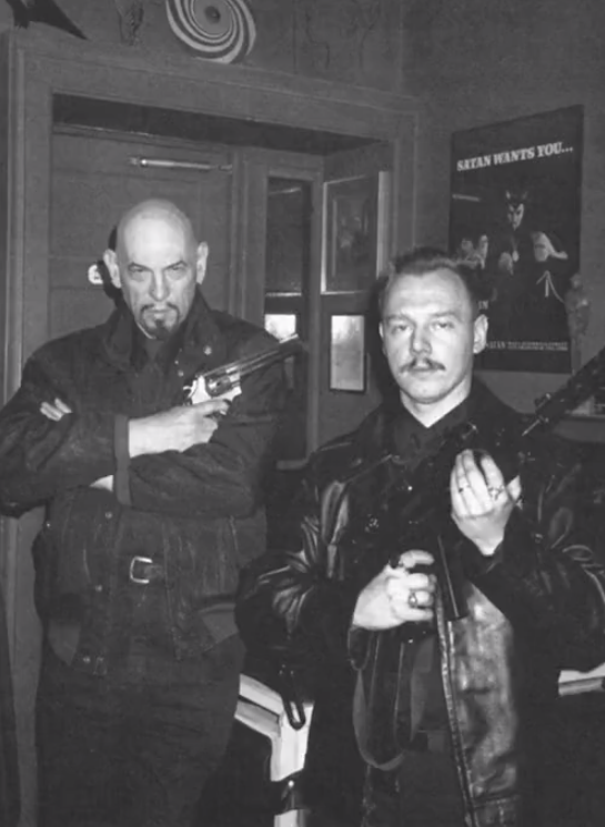 Black and white photo of Anton LaVey and Peter Gilmore standing, posing with a revolver and a rifle, respectively