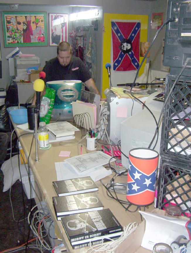 Early 2000s studio recording space with "Confederate flag" lamp cover on desk, "Might Is Right" books, and on wall, a Confederate flag with Charles Manson's face in the middle