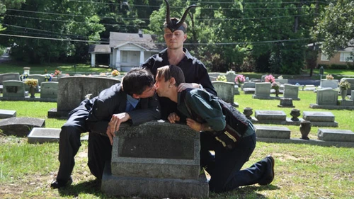 Gay couple kissing over the grave of Catherine Johnston with Doug Misicko in horned helmet as Lucien Greaves officiating in what was called a “pink mass”