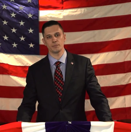 White male Augustus Sol Invictus stands at podium in a suit and tie with a USA flag behind him