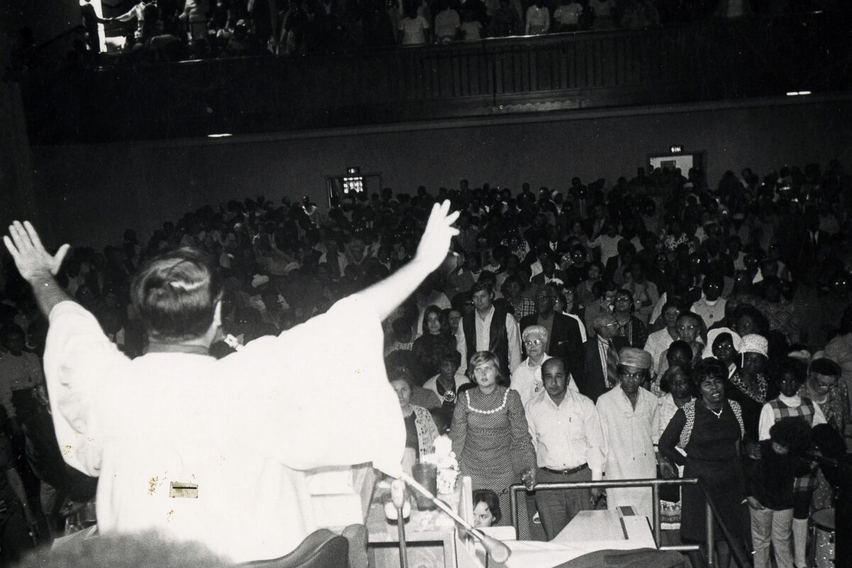 Image of Jim Jones from behind on a stage as he leads a service of The People's Temple, a large crowd in front of him