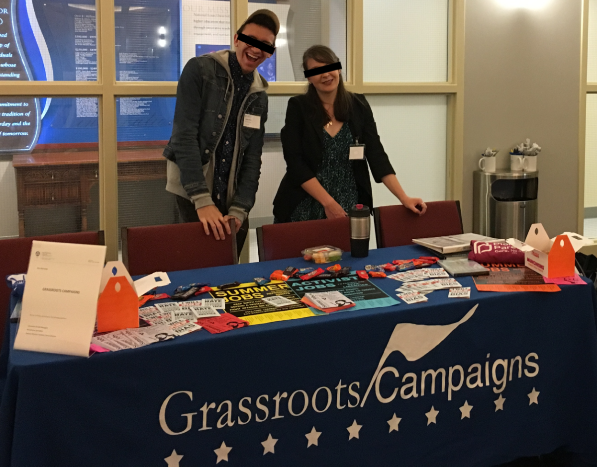 Two people with a black bars obscuring their eyes stand behind a Grassroots Campaigns recruitment table with pamphlets