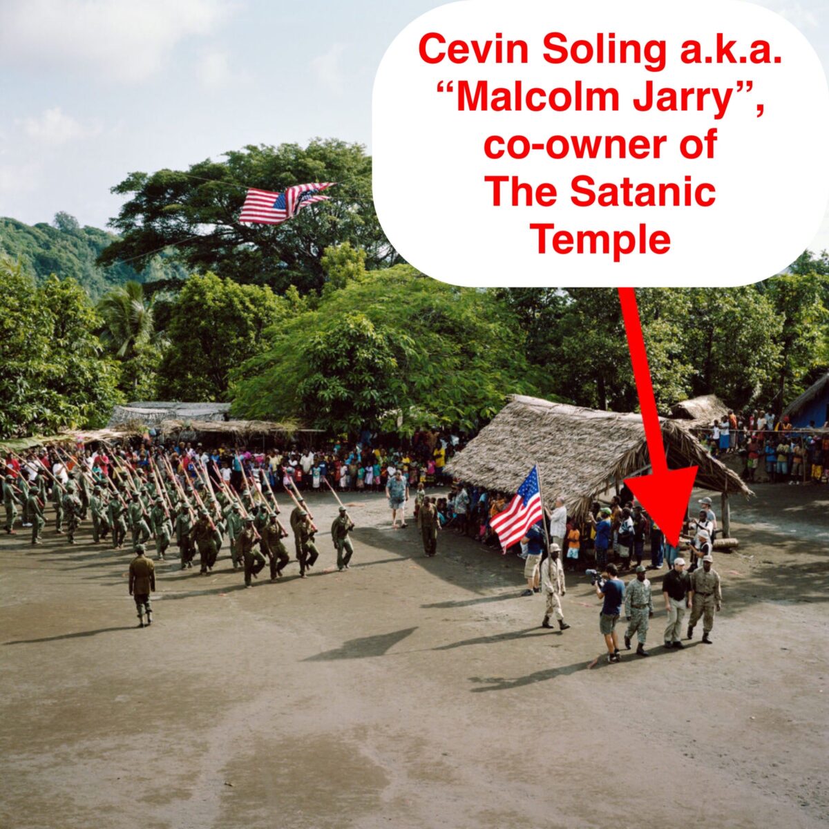 Arrow pointing at white man in khaki pant and a baseball cap, text says "Cevin Soling aka 'Malcolm Jarry', co-owner of The Satanic Temple." Soling is flanked by two Ni-Vanuatu men in mlitary fatigues, with another man carrying a USA flag a few steps behind him. Another six paces behind that man is a contingent of NI-Vanuatu men marching in fatigues with wooden muskets. From a hut and at the edge of a clearing, more people look on.