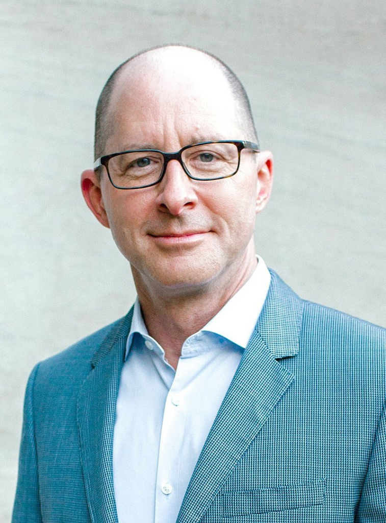 Photo of middle-agd bald white man in glasses wearing a suit with no tie, Charles R Eskridge III