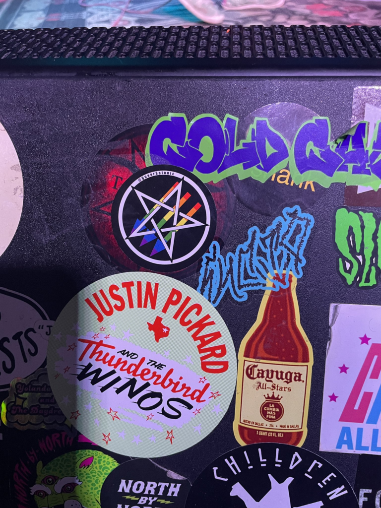 Slap sticker wall at a bar(?). Focus on a satanic antifascism sticker with gay pride colors by @QueerSatanic covering a sticker by The Satanic Temple underneath it