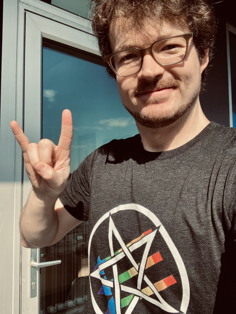 man in glasses throwing up the horns 🤘 while wearing a queer satanic antifascism shirt, white inverted pentacle over three arrows in gay pride colors