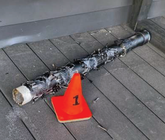 Photograph of pipebomb with orange evidence cone. "The IED appears to have been constructed from a section of plastic pipe covered with metal nails, which were attached to the pipe with tape. The inside of the pipe was filled with a powder-like substance, preliminarily
identified as smokeless gunpowder."