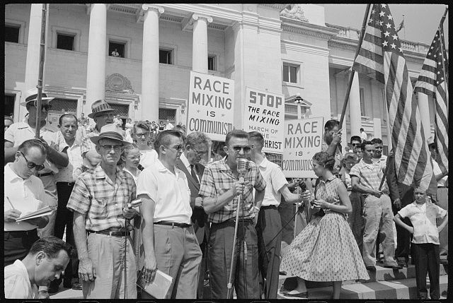 Man speaking at microphone in front of crowd at the Arkansas State Capitol protesting school integration, with signs reading "Race mixing is Communism" and "Stop the race mixing," Little Rock, Arkansas