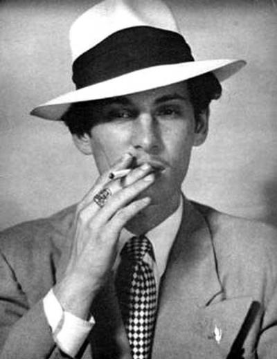 black and white photo of Anton LaVey as a young man, full on dressed like a gangster with the fedora and suit, smoking a cigarette.
